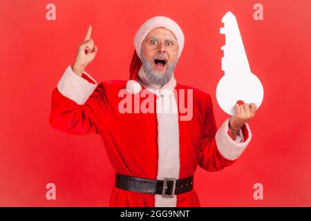 Porträt von staunenden älteren Mann mit grauem Bart trägt weihnachtsmann Kostüm stehen mit offenem Mund, hält Papierschlüssel, hat Idee für den Kauf von Immobilien. Stockfoto