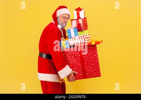 Porträt eines müden älteren Mannes mit grauem Bart, der das weihnachtsmann-Kostüm trägt und viele Schachteln mit Geschenken trägt, um sich auf Weihnachtsfeiern vorzubereiten. Ind Stockfoto