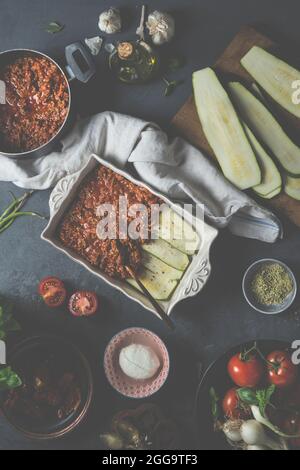 Zubereitung von Lasagne mit Zucchini und Bolognese-Sauce auf einem rustikalen dunklen Tisch mit verschiedenen Zutaten: Mozzarella-Käse, Zutaten, frisches Gemüse Stockfoto