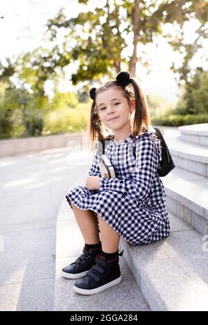 Nettes kleines Kind Mädchen 5-6 Jahre alt tragen karierte schwarz und weiß Kleid und Rucksack halten Bücher sitzen auf Treppen im Freien aus der Nähe. Der Camcorder wird angezeigt Stockfoto