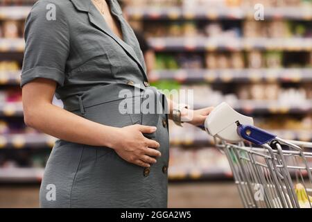 Nahaufnahme einer nicht erkennbaren Schwangeren in einem Kleid mit Gürtel, der die Hand am Bauch hält und den Wagen schiebt, während sie im Supermarkt Lebensmittel kauft Stockfoto