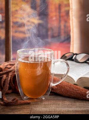 Dampfender heißer Tee in einem Glaskrug mit Decke und Buch an