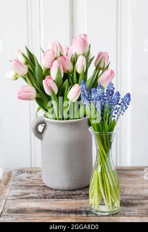 Bouquet von rosa Tulpen in grauer Keramikvase und Muscari-Blüten (Traubenhyazinthe) in Glasflasche. Holzhintergrund. Stockfoto