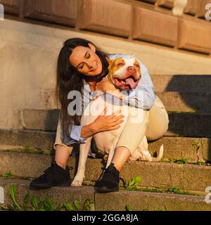 Frau, die auf einer Steintreppe sitzt und mit einem glücklichen amerikanischen Pitbull-Terrier kuschelt Stockfoto