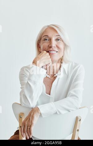 Vertikales Porträt einer eleganten, reifen Frau, die lächelt, während sie auf dem Stuhl rückwärts vor weißem Hintergrund sitzt Stockfoto