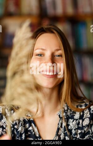 Ziemlich lächelnd freudig weiblich mit schönen Haaren, lässig gekleidet, mit Zufriedenheit in die Kamera schauen, glücklich sein. Aufnahme einer gut aussehenden schönen Frau an der Bücherregalwand. Stockfoto