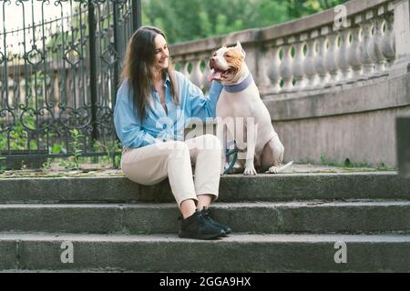 Frau, die auf einer Steintreppe sitzt und American Pitbull Terrier klopft. Glücklicher Hund, der den Besitzer ansieht Stockfoto