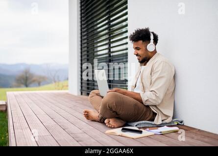 Junger Student mit Laptop und Kopfhörern, der zu Hause auf der Terrasse sitzt und studiert. Stockfoto