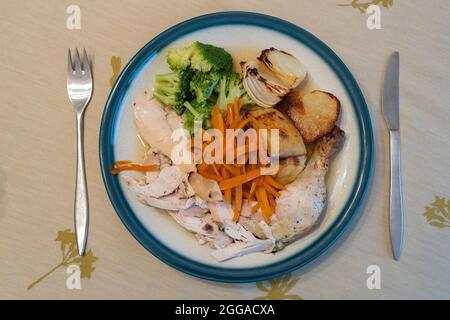 Ein vertikaler Blick auf ein typisch englisches Abendessen am Sonntag mit gebratenem Huhn, gebratenen Zwiebeln und Gemüse auf einem porzellanplatte mit Besteck Stockfoto