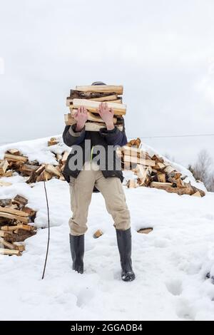 Der Mensch trägt an einem frostigen Wintertag eine Armladung gehacktes Holz in einem rustikalen Innenhof. Heizperiode im Dorf. Stockfoto