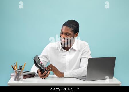 Erfolgreicher schwarzer Manager, der Rechner verwendet, während er am Arbeitsplatz sitzt, wobei der pc-Laptop auf blauem Hintergrund isoliert ist Stockfoto