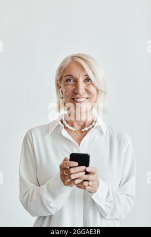 Vertikales Porträt einer eleganten, reifen Frau, die Smartphone hält und vor weißem Hintergrund die Kamera anlächelt Stockfoto