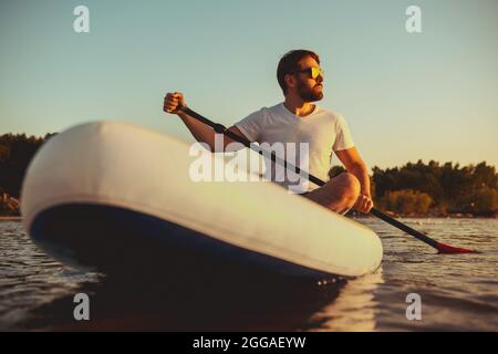 Hipster spaziert auf dem Boot am Sunset Lake. Paddle Boarder sitzt auf Paddleboards Stockfoto