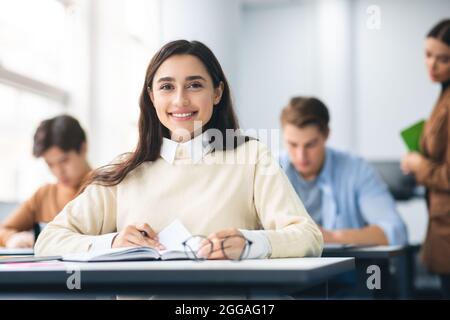 Lächelnder Student, der im Klassenzimmer am Schreibtisch sitzt und vor der Kamera posiert Stockfoto