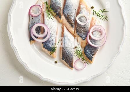 Gesalzener Hering mit Gewürzen, Kräutern und Zwiebeln auf weißem Teller auf hellem Steingrund mit Kopierfläche. Marinierter Fisch in Scheiben. Essen mit gesunder ungesättigter Nahrung Stockfoto