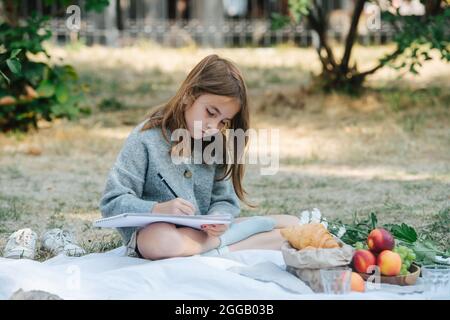 Kleines Mädchen, das auf einem Picknicktuch sitzt und Notizen auf einem Papier in einem Park schreibt. Sie zog ihre Schuhe aus, Obst und Croissants neben sich. Stockfoto