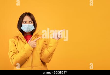 Junge Frau in Gesichtsmaske und Winterkleidung, die die Finger auf freien Raum über orangenen Studiohintergrund zeigt Stockfoto