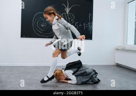 Schülerinnen spielen. Einer springt über den anderen zusammengerollt auf dem Boden. Tafel mit Zeichnungen im Hintergrund. Stockfoto