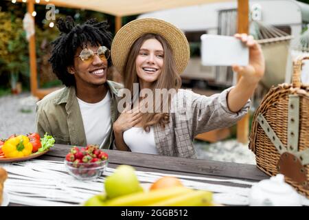 Kühle kaukasische Dame und ihr schwarzer Freund, die auf dem Campingplatz zu Mittag essen, Selfie in der Nähe des Wohnmobils machen und im Herbst im Freien ausruhen Stockfoto