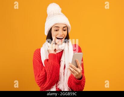 Abgelegenes Einkaufskonzept für Winterferien. Aufgeregt Frau in Strickmütze und Schal Blick auf Smartphone, gelber Hintergrund Stockfoto