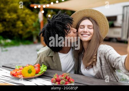 Liebevoller Afro-Kerl, der seine kaukasische Freundin küsst, während er im Herbst Selfie auf Campingausflug macht Stockfoto