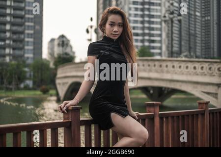 Schöne junge asiatische Frau in schwarzem Minikleid, die in der Nähe des Handlaufs im Park steht. Langes Haar. Schöne Haltung. Kamera mit Kopierplatz betrachten. Stockfoto