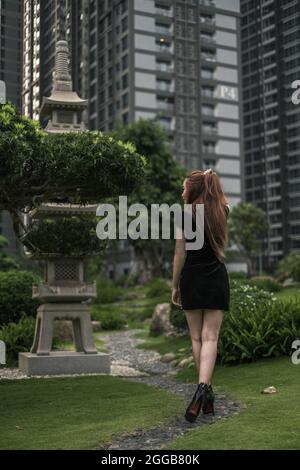 Schöne junge asiatische Frau in schwarzem Minikleid zu Fuß im Park. Langes Haar. Schöne Haltung. Rückansicht. Bild von hinten. Stockfoto