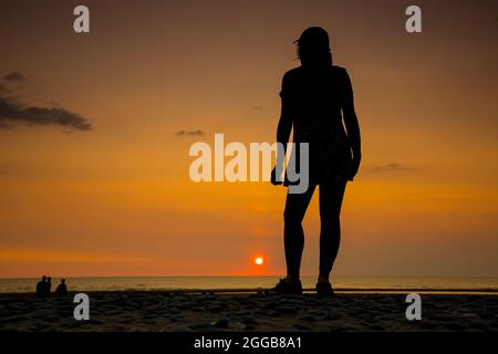 Rückansicht Silhouette einer isolierten jungen Frau, in Sportmütze, Blick auf das Meer bei Sonnenuntergang auf einem britischen Strand, Aufenthalt Sommerurlaub. Stockfoto