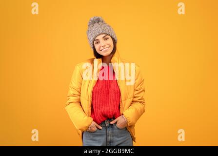 Fröhliche Dame in warmer Jacke, Strickpullover und Hut, die Hände in den Taschen hält und vor der Kamera lächelt, orangefarbener Hintergrund Stockfoto