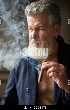 Porträt eines brutalen grauhaarigen reifen Mannes, der wegschaut, während er Zigarre hält Stockfoto