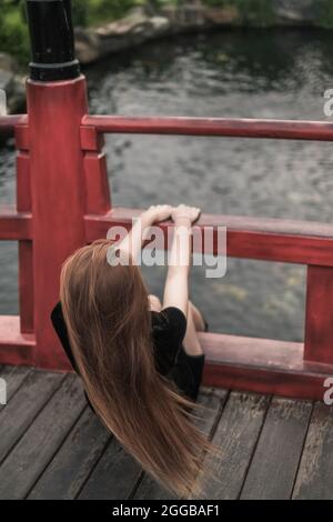 Rückseitenansicht Bild einer jungen Frau in lässigem schwarzen Kleid, die im Park auf der Brücke mit Handläufen sitzt. Lange schöne Haare Stockfoto