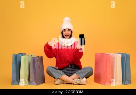 Aufgeregt Frau im Winter Hut Online-Shopping auf Smartphone sitzt zwischen bunten Papiertaschen auf gelbem Hintergrund Stockfoto