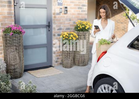 Frau mit Telefon in der Nähe des Autos auf der Veranda ihres Hauses Stockfoto