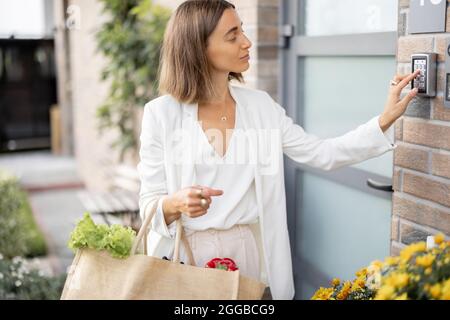 Frau kommt mit Lebensmitteln nach Hause und gibt einen Code ein, um die Tür zu betreten Stockfoto