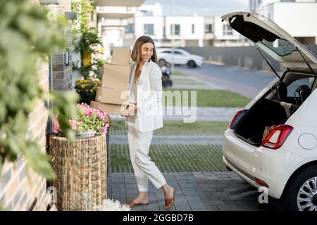 Junge Geschäftsfrau, die Pakete von einem Kofferraum abholt und mit dem Auto nach Hause kommt. Konzept, Waren online zu kaufen und nach Hause zu liefern Stockfoto