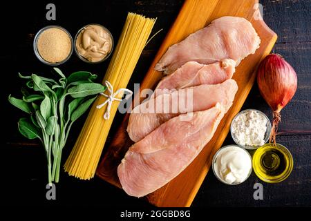 Cremiges Senfhuhn mit Engelshaar-Pasta Zutaten: Chicken Cutlets, Capellini, frischer Salbei und andere Zutaten auf einem dunklen Holztisch Stockfoto