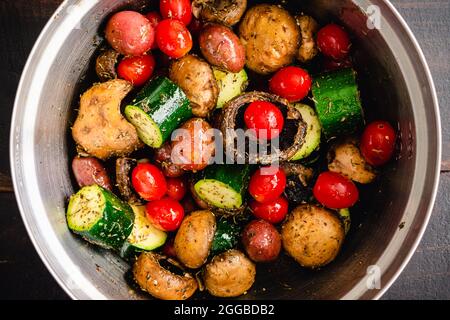 Ungekochte italienische Gemüsesorten in Olivenöl und Gewürzen: Rohe Traubentomaten, Zucchini, Pilze, rote Kartoffeln, Knoblauchzehen in einer Rührschüssel Stockfoto