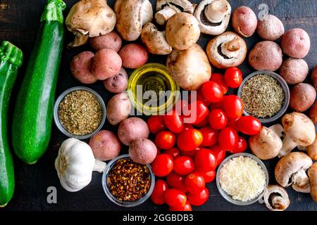 Italienische Ofen geröstetes Gemüse Zutaten auf einem Holztisch: Traubentomaten, Zucchini, Pilze, rote Kartoffeln, Kräuter, Gewürze und Olivenöl Stockfoto