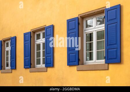 Drei weiße Fenster mit Sandsteinschwellen in einer Reihe mit dunkelblauen Fensterläden und einer orangegelben Hauswand Stockfoto