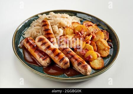 Teller mit gegrillten würzigen Würstchen, Kohl und knusprigen Kartoffelscheiben mit Speckstückchen, gekrönt mit einer reichen Soße und serviert auf weißem Hintergrund in einem hohen Stockfoto