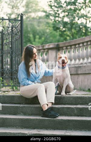 Frau, die auf einer Steintreppe sitzt und American Pitbull Terrier streichelt. Glücklicher Hund, der die Kamera anschaut Stockfoto
