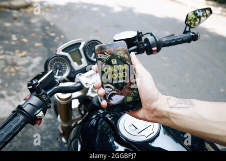 Hand eines Mannes, der auf dem Motorrad sitzt und die Anwendung der Lebensmittelzustellung auf dem Smartphone zeigt Stockfoto