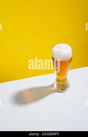 Kaltes Bier in einem Glas mit weißem Schaum auf einem weißen Tisch und gelbem Hintergrund serviert, keine Menschen Stockfoto