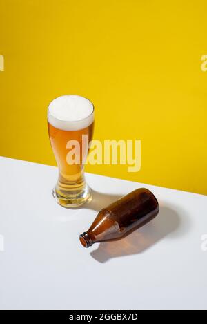 Kaltes Bier serviert in einem Glas mit weißem Schaum, eine leere Glasflasche auf einer Seite, auf einem weißen Tisch und gelbem Hintergrund, keine Menschen Stockfoto