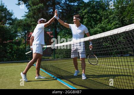 Lächelnde, multiethnische Spieler mit Tennisschlägern, die auf dem Platz hohe fünf geben Stockfoto