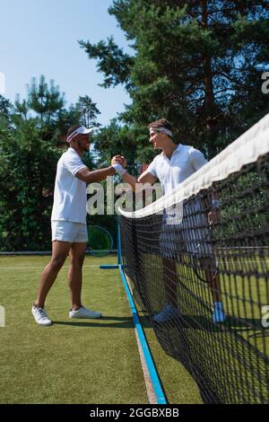 Seitenansicht von fröhlichen, interracial Tennisspielern, die sich auf dem Platz in der Nähe des Netzes die Hände schütteln Stockfoto