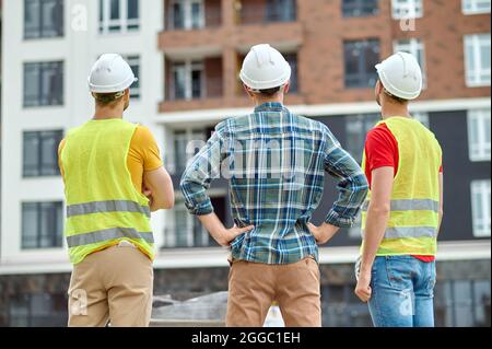 Drei Arbeiter in Harthüten stehen vor einem neuen Gebäude Stockfoto