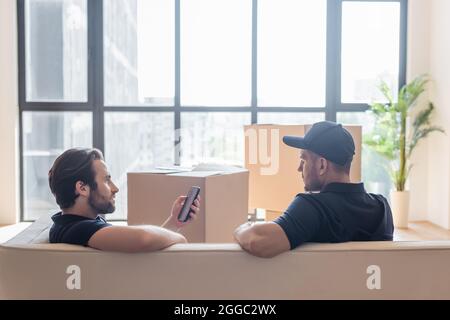 Arbeiter mit Smartphone auf dem Sofa in der Nähe von Kollegen und Kartonverpackungen Stockfoto