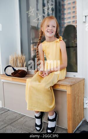 Sommersprossen-Ingwer-Mädchen in einem lebendigen gelben Kleid sitzt auf einer Bank. Füße hängen in der Luft und schauen auf die Seite. Stockfoto