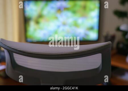 Blick über die Kopfstütze eines modernen ergonomischen Bürostuhls auf einen hell erleuchteten LCD-Bildschirm mit Breitbild-Bildschirm im Home-Office Stockfoto